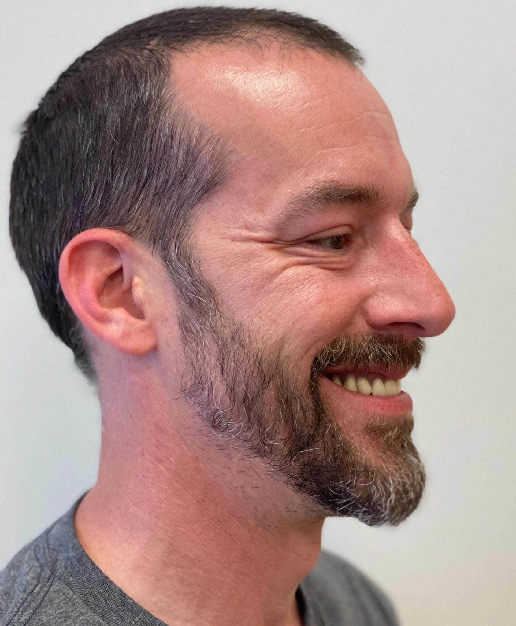 The image shows a man with a beard and mustache, smiling slightly, against a neutral background.