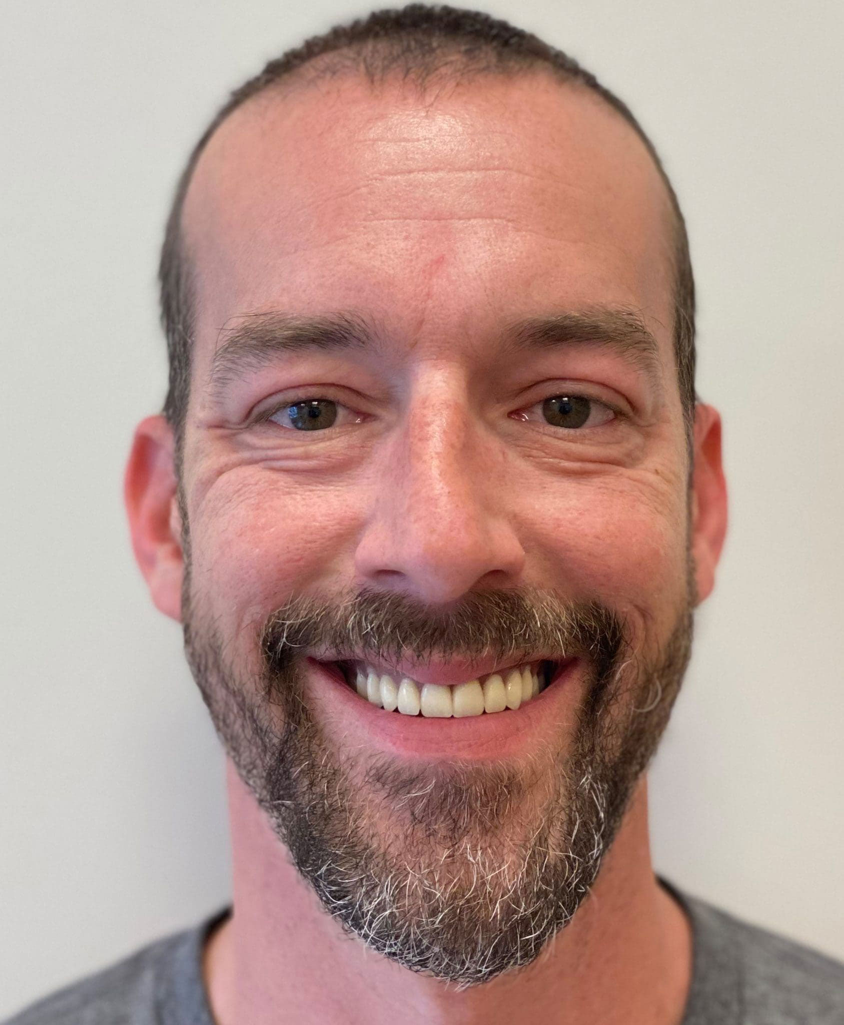 The image shows a man with a beard and mustache, smiling at the camera. He has short hair and appears to be indoors with a plain background.