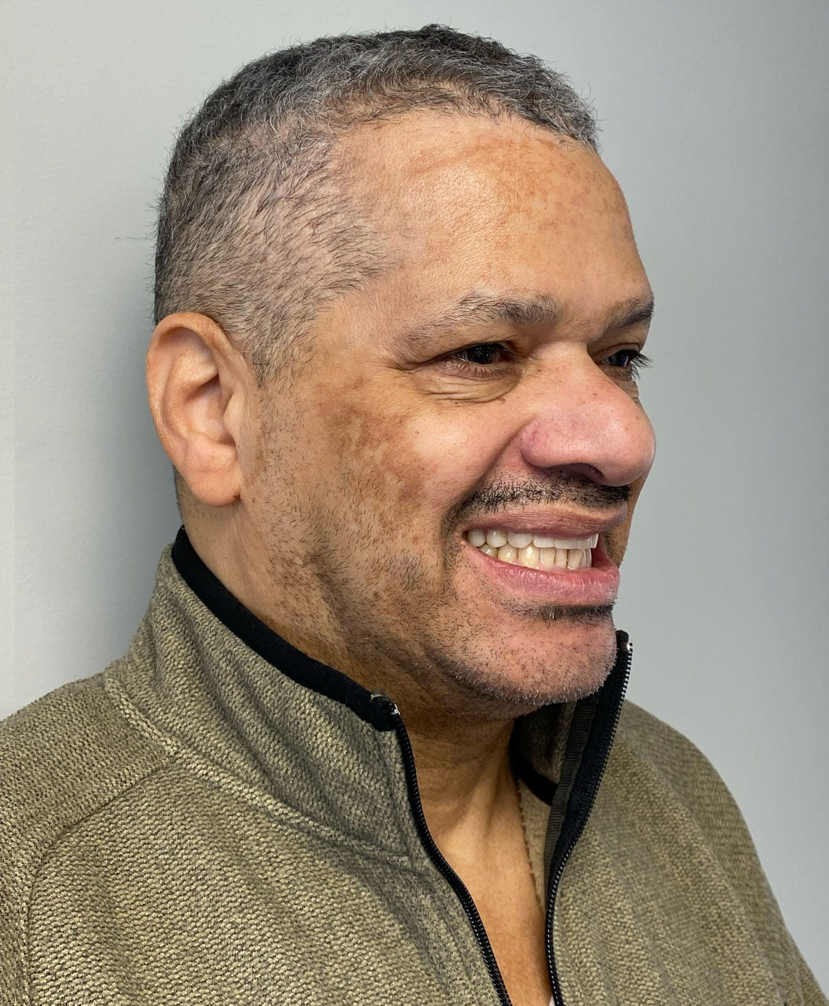 The image shows a man with a mustache, smiling and looking directly at the camera. He has a bald head and is wearing a dark-colored jacket.