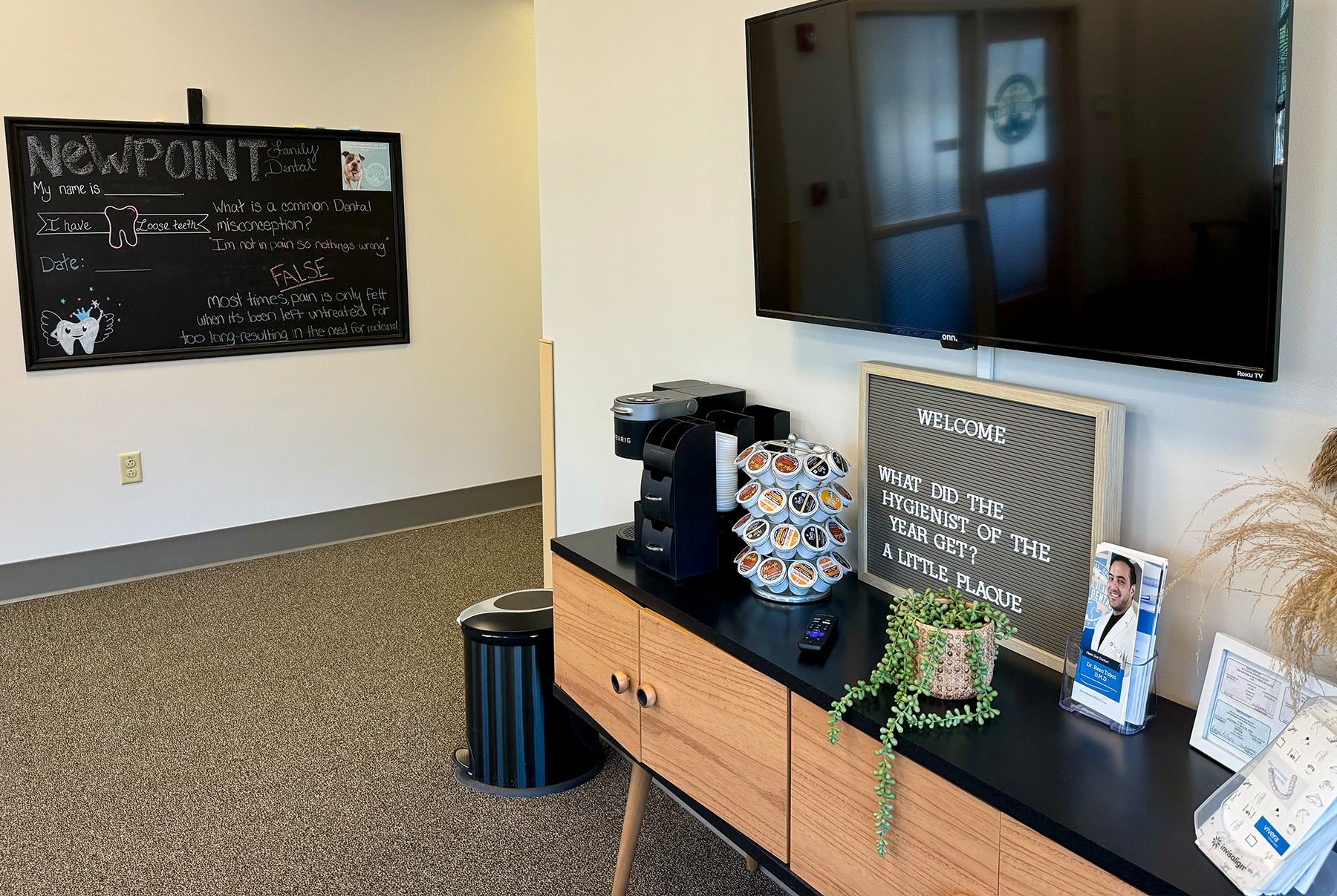 The image shows an office reception area with a television screen displaying the channel logo, a coffee station, a sign with a message, and a framed photo on a desk.