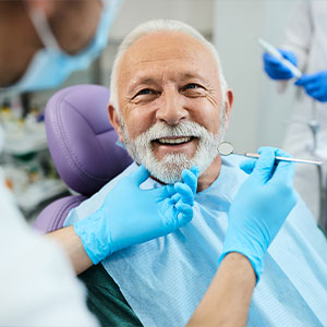 The image depicts an older man with a beard and mustache receiving dental care, seated in a dental chair while a dental professional works on him.