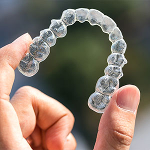 The image shows a person s hand holding a clear, transparent dental retainer that is shaped like a smile.