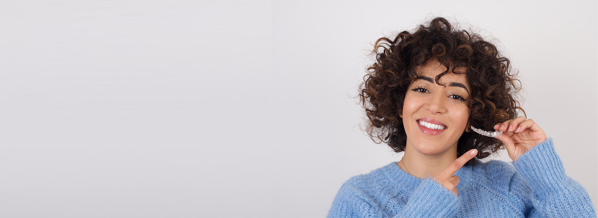 A woman with short curly hair is smiling and holding a smartphone, standing against a neutral background.