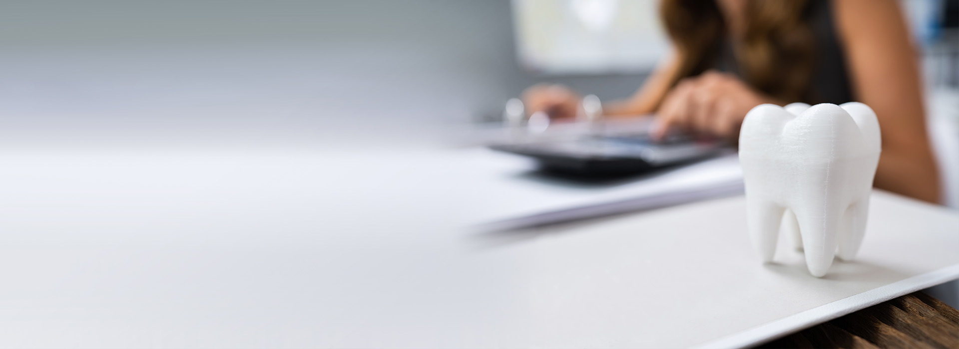 The image shows a person sitting at a desk with a computer, working on a document. In the foreground, there is a 3D model of a tooth, and in the background, there s a blurred view of a person using a laptop.