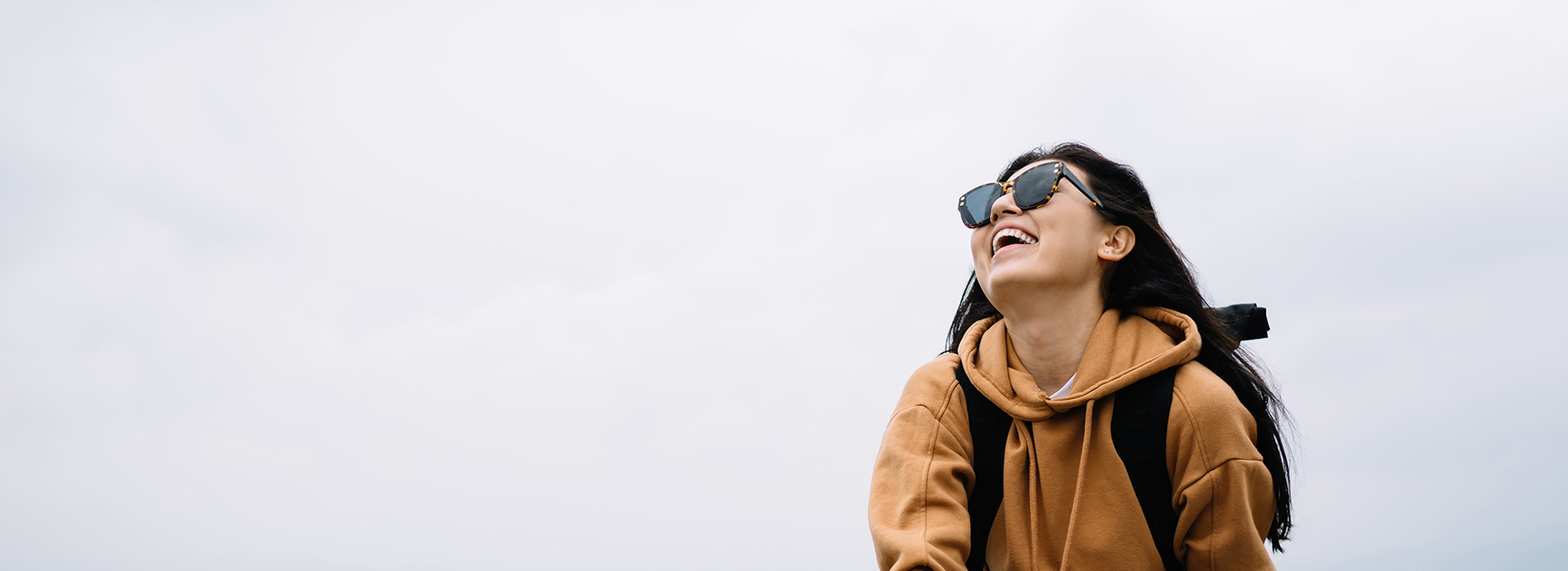 The image shows a person standing outdoors, smiling towards the camera, with sunglasses and a hoodie, against a backdrop of an overcast sky.