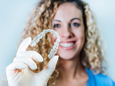 A woman holds up a clear dental retainer with her right hand, smiling at the camera.