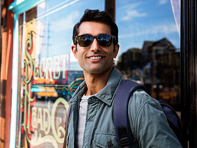 A man stands confidently in front of a storefront, dressed casually with sunglasses and a backpack.