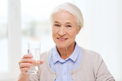 The image shows an elderly woman holding a glass of water and smiling, with a clear background.