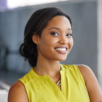 A woman with an open smile, wearing a yellow top and standing in front of a building.