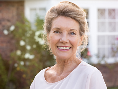 The image is a photograph of an elderly woman smiling at the camera. She has short blonde hair, wears makeup with prominent red lipstick, and is dressed in a white top with a light-colored cardigan. Her background features a house with a brick exterior and a well-maintained garden with a variety of plants and shrubs.