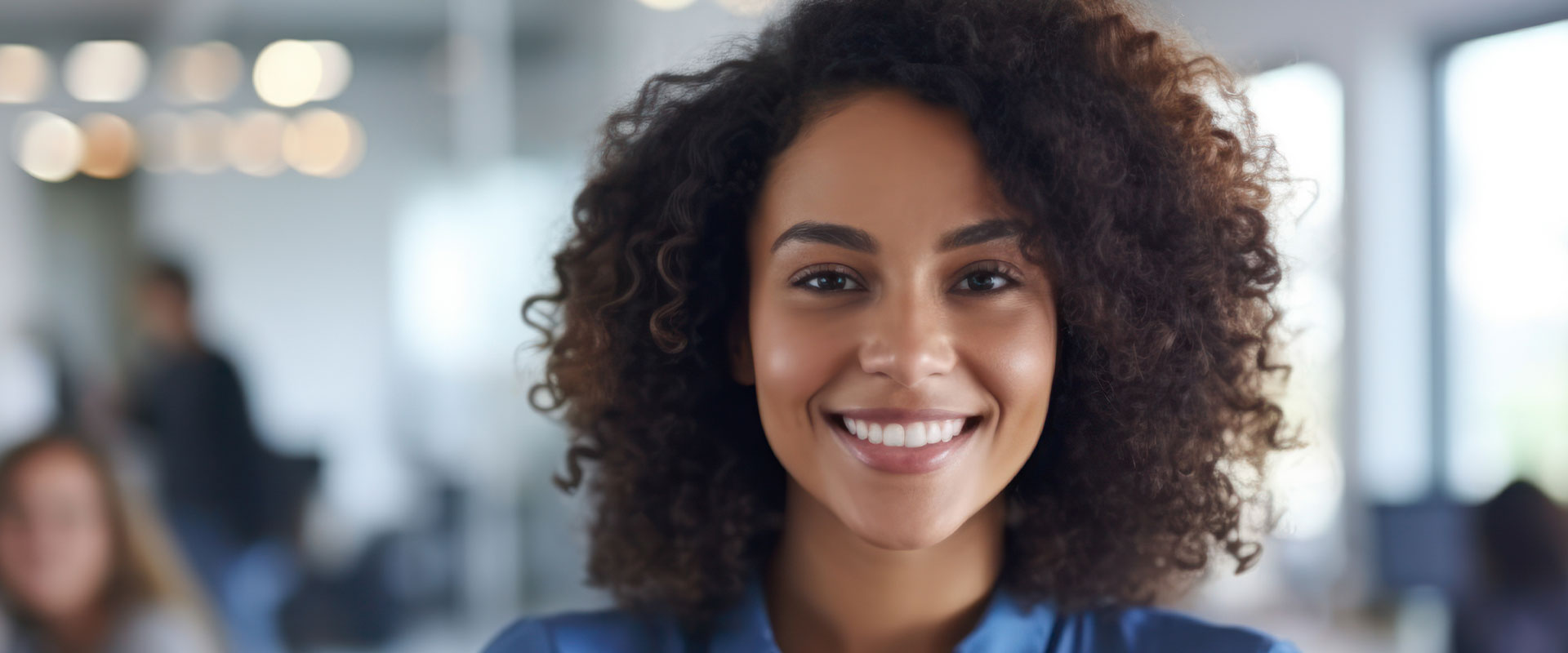 The image depicts a smiling individual with curly hair, wearing a blue top, standing in an office environment.