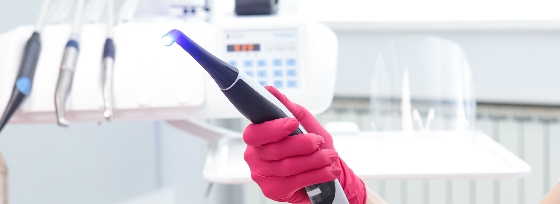 A person in a pink glove holding a blue device, possibly a stylus or pen, near a white surface with various objects and a blurred background.