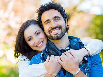 A man and woman hugging each other, smiling, with the man wearing a scarf around his neck.