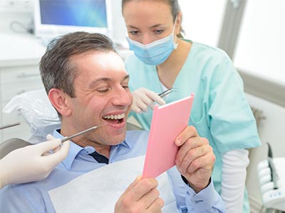 The image depicts a man in a dental chair, holding up a pink card or paper with a smile on his face, while a dentist and dental hygienist are present in the background.