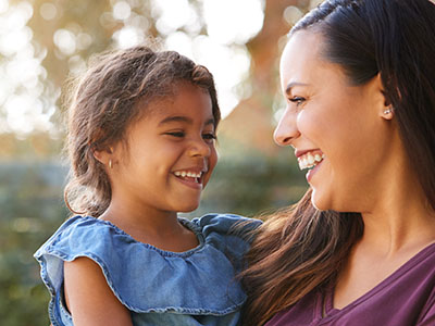 The image shows a woman and a young child, both smiling, with the woman holding the child. They appear to be outdoors during daylight.