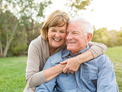 This is a color photograph featuring an older couple embracing each other outdoors. The man is wearing a blue shirt and the woman has blonde hair, both are smiling and appear to be in their senior years. They are standing on grass with trees visible in the background.