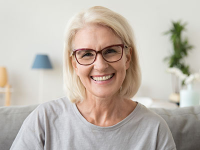 The image is a photograph of an older woman with short blonde hair, wearing glasses and a light-colored top. She is smiling at the camera and appears to be indoors with a domestic setting in the background.