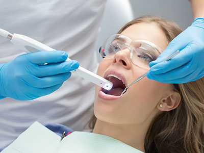 The image shows a dental professional using a device to clean or examine a patient s teeth while the patient is seated in a dental chair, wearing protective eyewear.