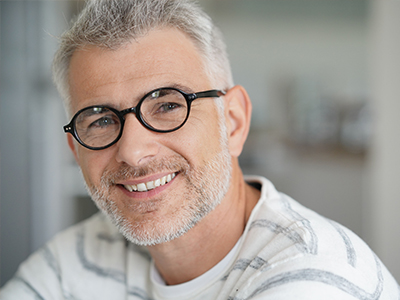 The image shows a man with gray hair, wearing glasses and a white shirt, smiling at the camera. He appears to be in an indoor setting, possibly an office or a professional environment.