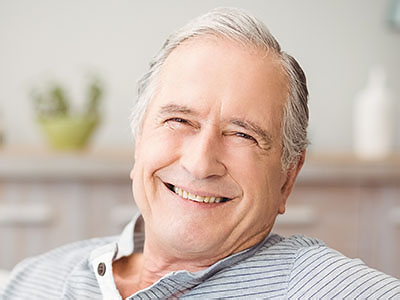 A man with gray hair, smiling and wearing a blue shirt, seated in front of a plant.