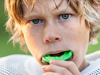 The image is a photograph of a young male athlete with fair skin, wearing a white football uniform and sporting blonde hair. He appears to be holding a green mouthguard in his hand and has an intense expression on his face.